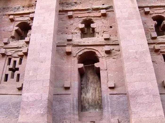 A close-up view of a stone building showcasing its architectural details, including arches and weathered textures, set against a clear outdoor background. The structure appears to be part of an ancient historical site or ruins.
