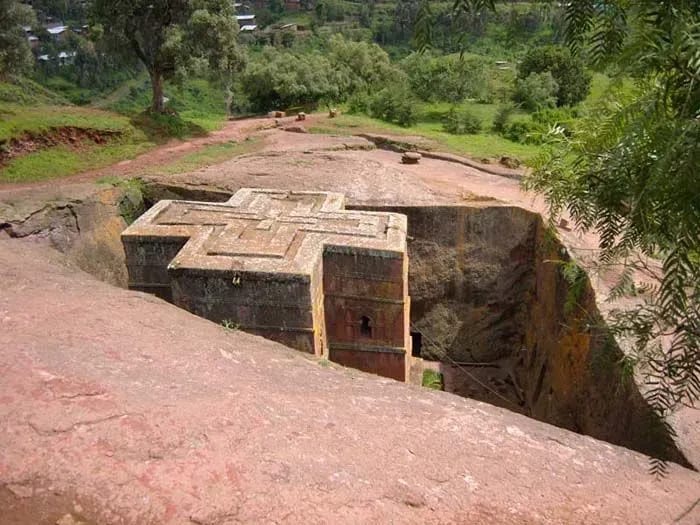 A stone building situated inside a cave with the landscape of Lalibela in the background, surrounded by trees and rocky terrain.